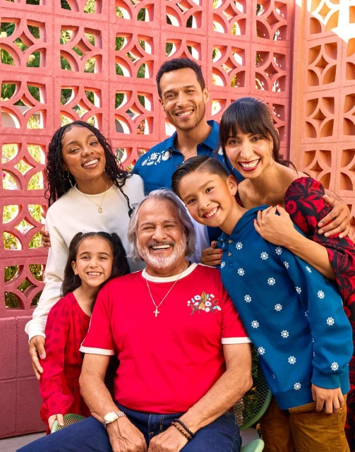 Group portrait of a multigenerational family smiling outdoors against a coral-colored patterned wall, creating a warm and joyful atmosphere.