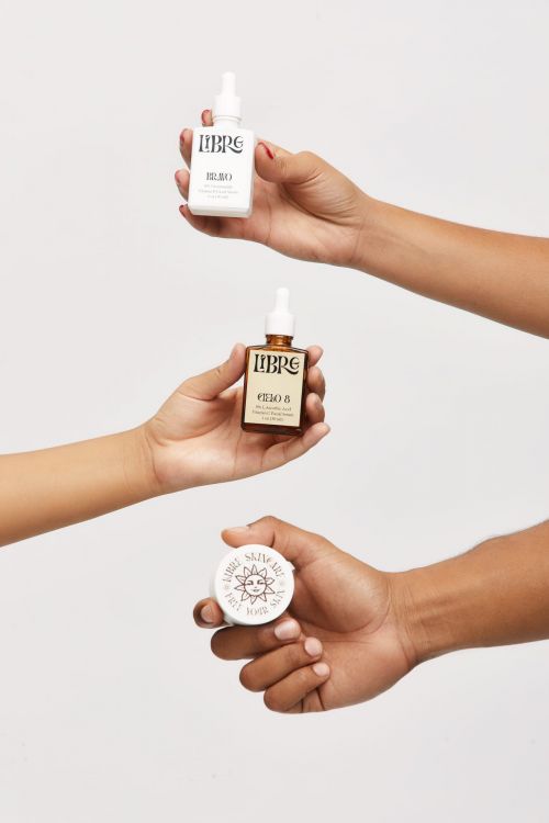 Three hands present a white and brown serum bottle plus moisturizer against a white backdrop in a beauty, skincare product shot.