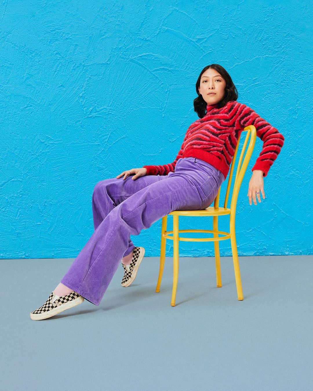 A young adult woman with dark hair poses in a red patterned top and purple pants, seated on a yellow chair against a blue textured wall.