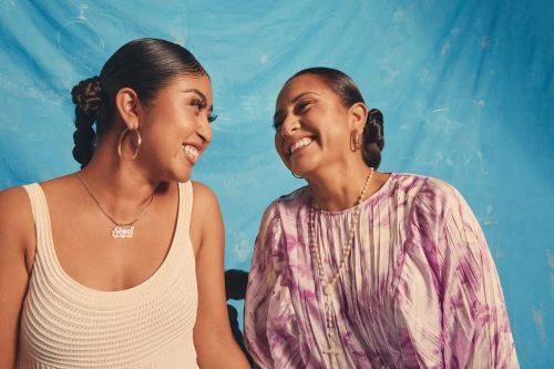 Portrait of a smiling mother and daughter posing indoors against a blue backdrop, wearing light-colored clothing and jewelry.