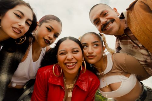 A group of five young adults, including a male, pose closely together looking down at the camera with bright smiles outdoors in daylight.
