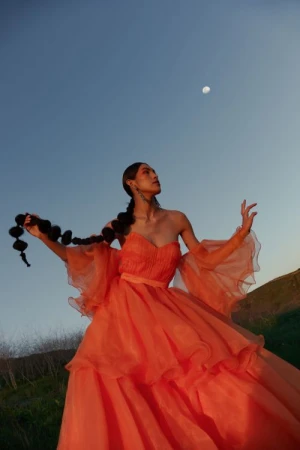 Woman in a vibrant orange strapless gown holding a long braided hairpiece beneath a clear twilight sky with a visible moon.