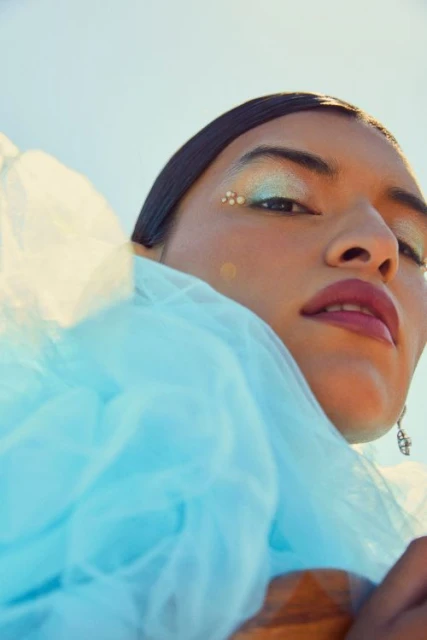 Eye-level shot of woman with glitter eyeshadow, pearl embellishments, and blue tulle fabric against a bright, light-blue backdrop.