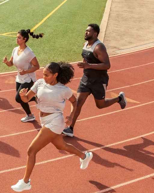Three athletes run on a red track; the foremost runner is a woman with dark skin in gray athletic wear and white running shoes.
