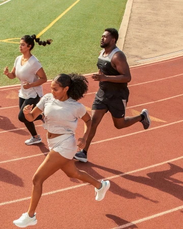 Three athletes run on a red track; the foremost runner is a woman with dark skin in gray athletic wear and white running shoes.