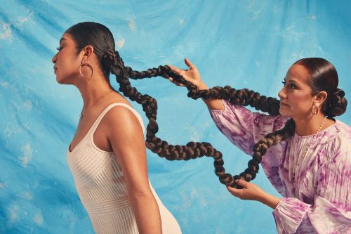 Two women with long braids: one with profile view in white dress, and another holding the braid, on a blue painted background.