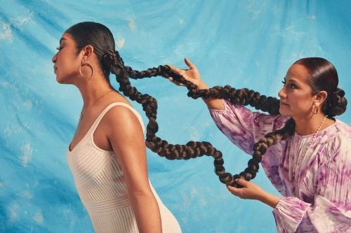 Two women with long braids: one with profile view in white dress, and another holding the braid, on a blue painted background.