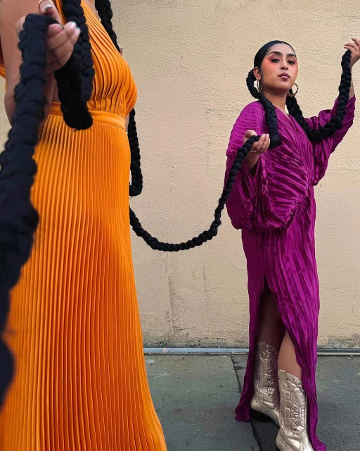 Two women in pleated dresses pose outdoors, holding long braids. One dress is orange, the other is fuchsia; both are styled with cowboy boots.