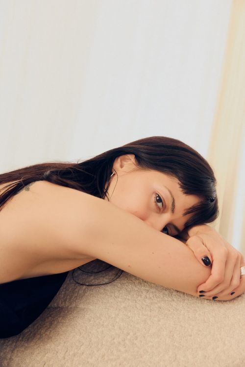 Close-up portrait of a brunette woman with bangs resting her head on her arms indoors, illuminated by soft natural light, creating a serene mood.