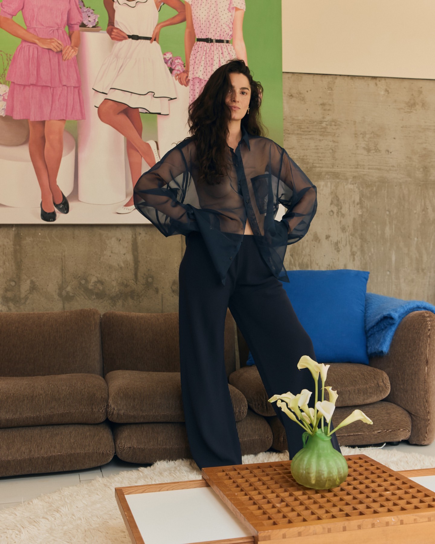 A woman in a sheer blouse and pants poses in an indoor setting with a painting and a couch in the background, creating a stylish atmosphere.