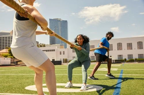 Group of three people, two women and a man, engaged in a tug-of-war on a sunny outdoor field with buildings and blue sky in background.