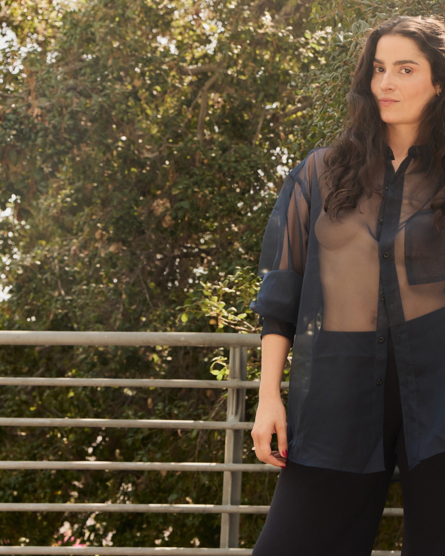 Portrait of an adult woman with long dark hair wearing a sheer blouse standing on a balcony with trees in the background.