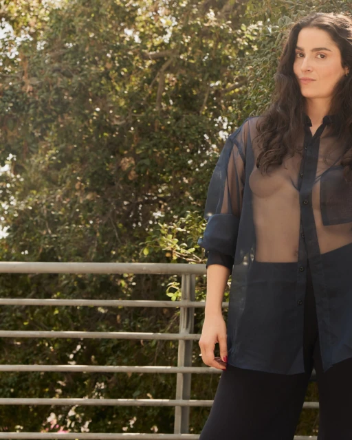 Portrait of an adult woman with long dark hair wearing a sheer blouse standing on a balcony with trees in the background.