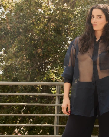 Portrait of an adult woman with long dark hair wearing a sheer blouse standing on a balcony with trees in the background.