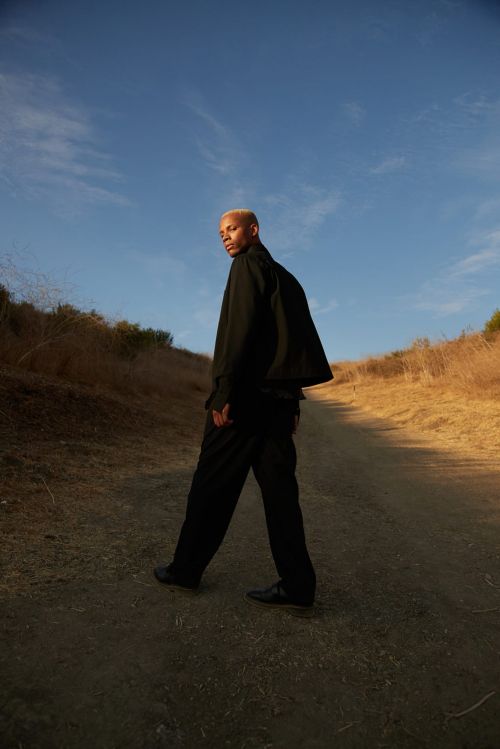 An adult Black male with blond hair walks on a dirt path outdoors wearing black apparel, against a background of blue sky and dry vegetation.