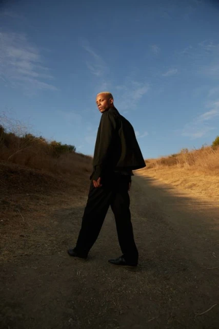 An adult Black male with blond hair walks on a dirt path outdoors wearing black apparel, against a background of blue sky and dry vegetation.
