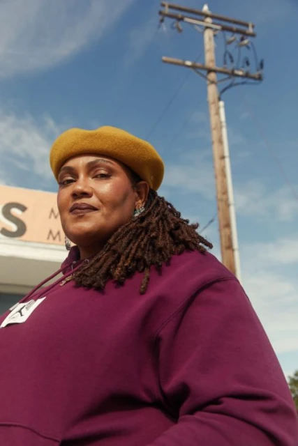 Portrait of an adult woman wearing a beret and burgundy hoodie in an outdoor urban environment with a utility pole.
