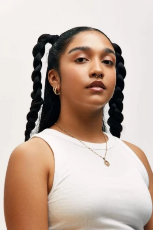 Portrait of a young woman with braided pigtails wearing a white tank top and gold hoop earrings against a white background