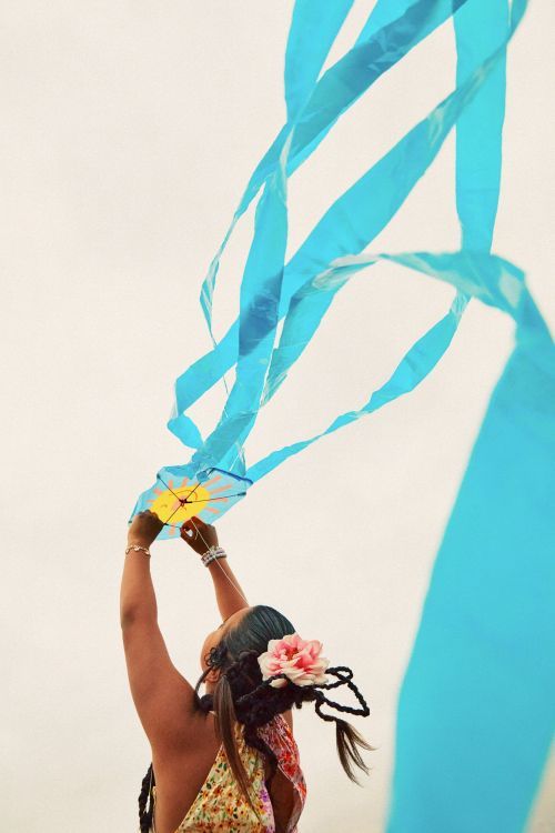 Young woman from behind with a floral hairpiece holds up a kite with long blue streamers against a light sky in a vertical composition.