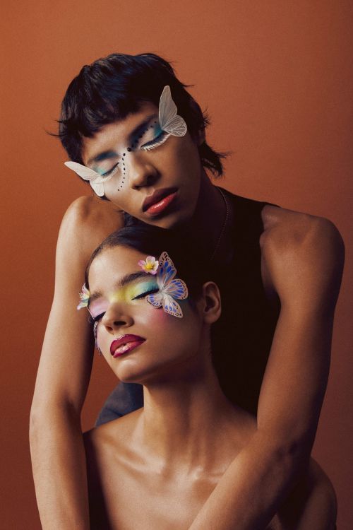 Two women pose closely together with creative makeup, including butterfly accents on their faces, against a plain brown background.