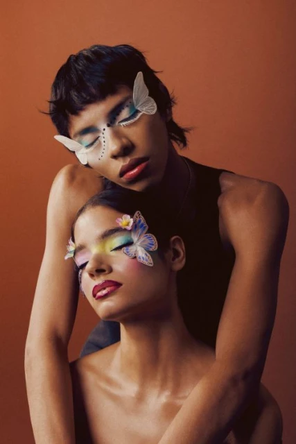 Two women pose closely together with creative makeup, including butterfly accents on their faces, against a plain brown background.