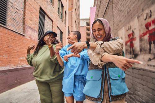 Three smiling women of diverse ethnicity pose in street style against an urban alleyway backdrop in a candid, eye-level, medium shot.