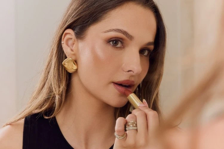A woman applies lipstick in front of a mirror, showcasing her gold earrings, multiple rings, and dark sleeveless top in an indoor setting.