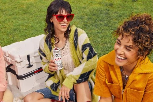 Two young women laugh together on the grass with a cooler and a can of hard seltzer, wearing casual clothing, in an outdoor setting.