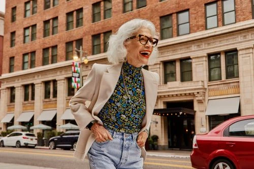 Elderly woman smiles, wearing glasses and a blazer, walking on a city street with brick buildings and parked cars in the background.