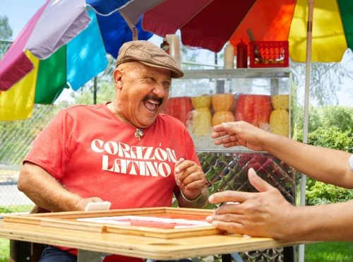 Enthusiastic adult male playing dominoes outdoors at a street food cart, wearing a red Corazon Latino t-shirt and newsboy cap.