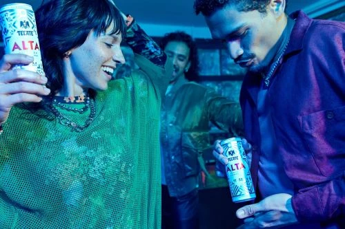 Three people are in a dimly lit room, two adult males and one adult female, holding canned drinks. The scene has a party atmosphere.