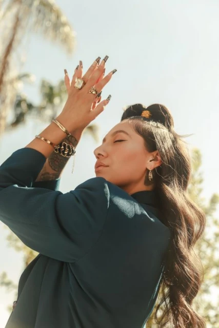 Woman in a blue blazer poses with her hands raised toward the sky, showcasing rings and bracelets against a blurred outdoor background.