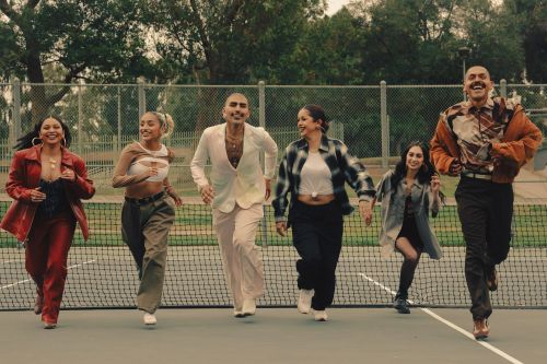 Six adults in fashionable casual wear run toward the camera outdoors on a tennis court with greenery and overcast sky in the background.