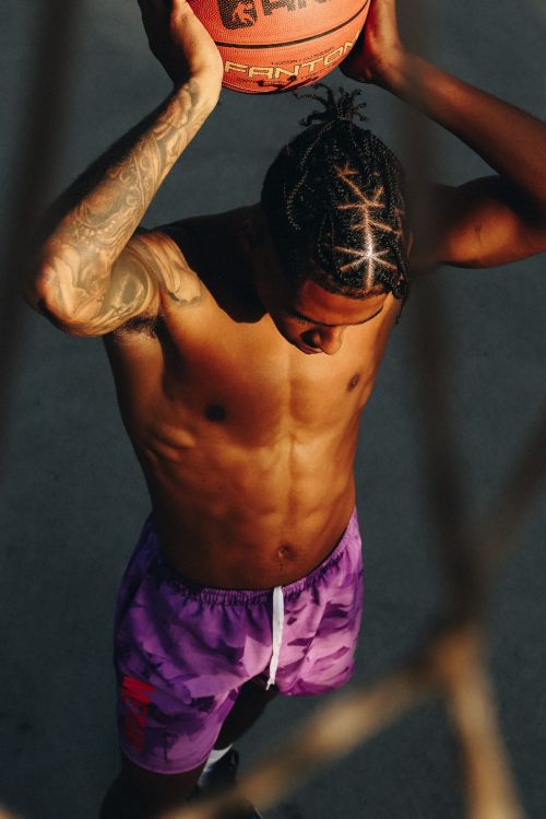 Top-down shot of an adult male athlete with tattoos holding a basketball over his head outdoors; he wears purple camouflage shorts.