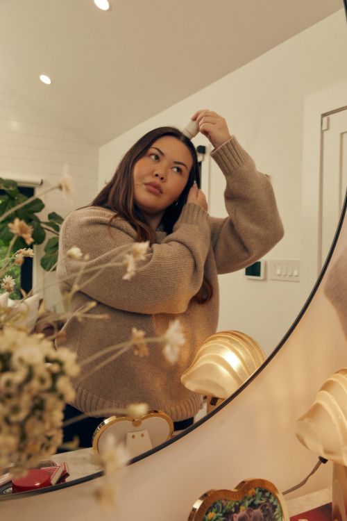 An adult female is applying powder to her face in the bathroom mirror. The scene features soft, natural lighting, home interior setting, and beauty product.