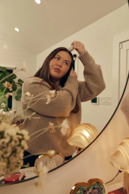 An adult female is applying powder to her face in the bathroom mirror. The scene features soft, natural lighting, home interior setting, and beauty product.