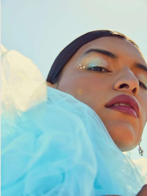 Close-up portrait of a woman with iridescent makeup and a light blue ruffled garment against a clear, bright blue sky background.