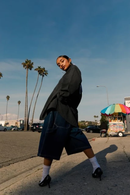 Stylish woman posing outdoors in oversized blazer and denim shorts with palm trees and blue sky in the background during daytime.