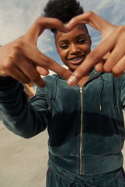 A smiling Black woman makes a heart shape with her hands in an outdoor setting. She is wearing a velour tracksuit with braces.
