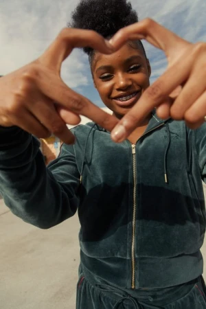 A smiling Black woman makes a heart shape with her hands in an outdoor setting. She is wearing a velour tracksuit with braces.