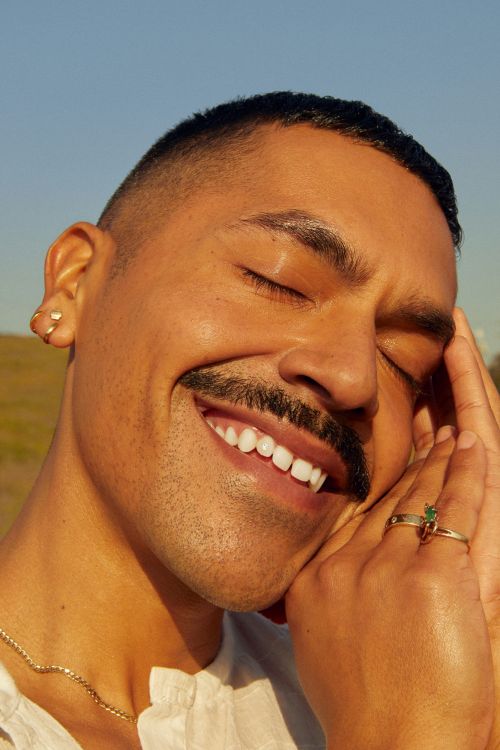Close-up of a smiling adult male with a mustache, eyes closed, and hands gently resting on his face, set against a clear blue sky.