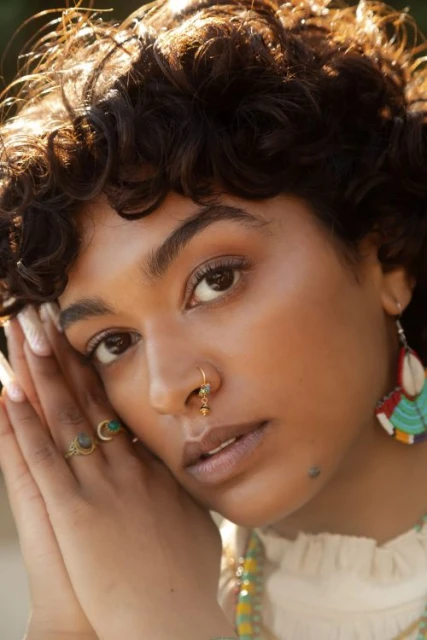 Close-up portrait of a woman with dark curly hair, fair skin, and a nose piercing, posing outdoors in soft light.