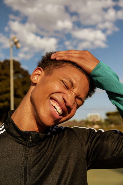 Young man in a black tracksuit with white stripes smiles and poses outdoors in a sports setting on a sunny day with a blue sky.