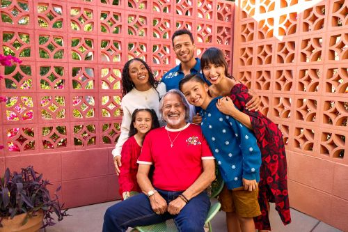 Family portrait of six people smiling outdoors in front of a decorative pink wall with lush greenery, creating a warm, inviting atmosphere.