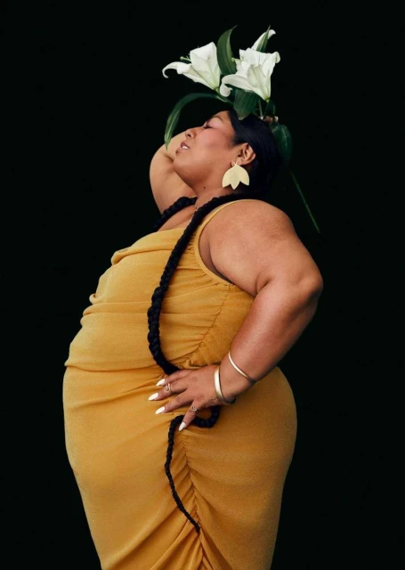 Portrait of a plus-size woman in a yellow dress with white flowers, a black necklace, and gold jewelry against a black backdrop.