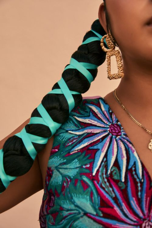 Close-up shot of an adult woman with braided hair wrapped in turquoise ribbons, gold earrings, and a flower-patterned top.