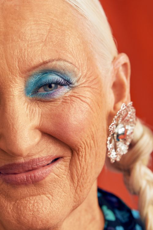Close-up of an elderly woman with white hair, wearing vivid blue eyeshadow and an elaborate silver earring. Orange background.