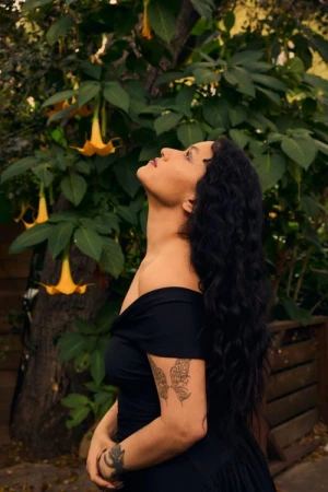 Profile portrait of a woman with long dark wavy hair in a black dress looking up outdoors near hanging yellow flowers.
