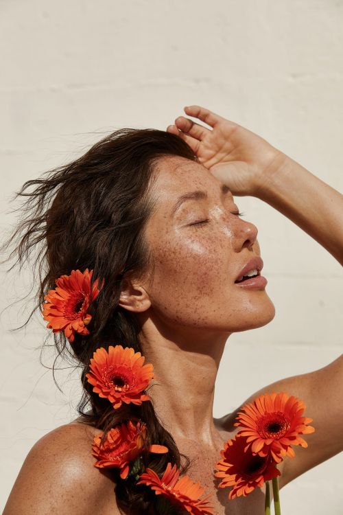 Woman with Gerbera daisies braided in her dark hair, touching her forehead, in soft light against a white background, beauty shot.