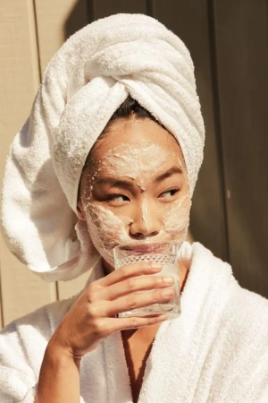 A woman with a white towel wrapped around her head and a white bathrobe holds a glass of water, a face mask is applied.